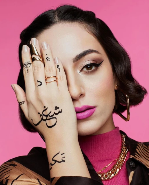 Close-up shot of a woman with henna tattoos on her hand, statement rings, pink lipstick, and a pink background for a beauty ad.
