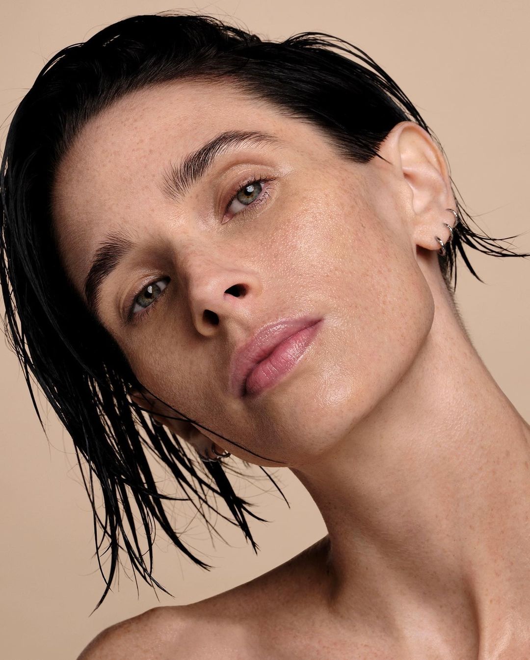 Close-up beauty shot of a fair-skinned woman with dark, slicked-back hair and neutral makeup posing against a beige background in portrait format.