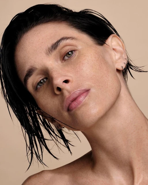 Close-up beauty shot of a fair-skinned woman with dark, slicked-back hair and neutral makeup posing against a beige background in portrait format.