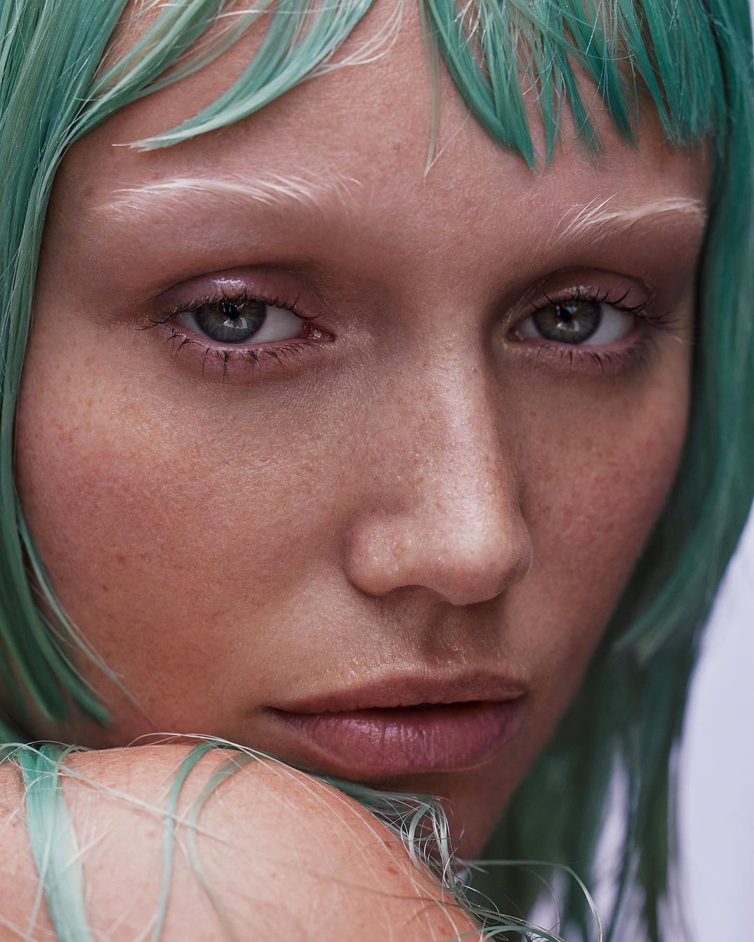 Close-up portrait of a fair-skinned woman with grey eyes, green tinted hair, and light eyebrows looking directly at the camera.