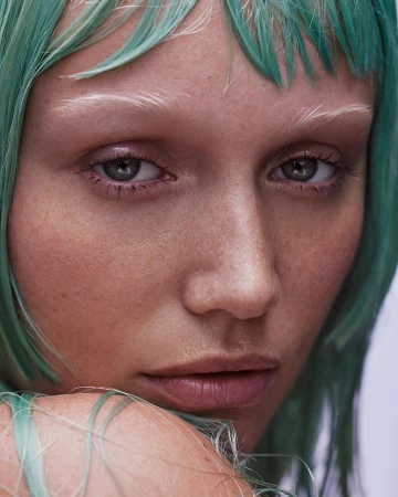 Close-up portrait of a fair-skinned woman with grey eyes, green tinted hair, and light eyebrows looking directly at the camera.