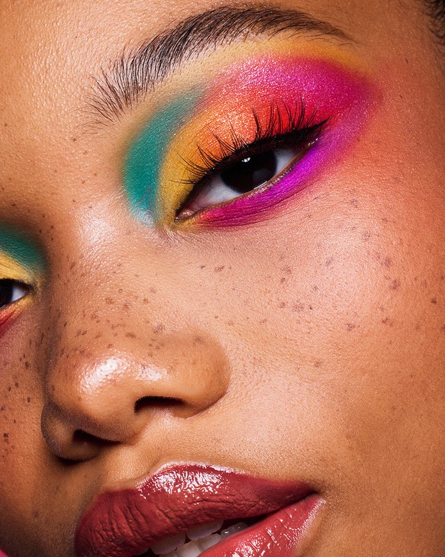 Close-up of a person's face showcasing vibrant rainbow eye makeup. The image highlights the colorful eyeshadow and freckled skin texture.