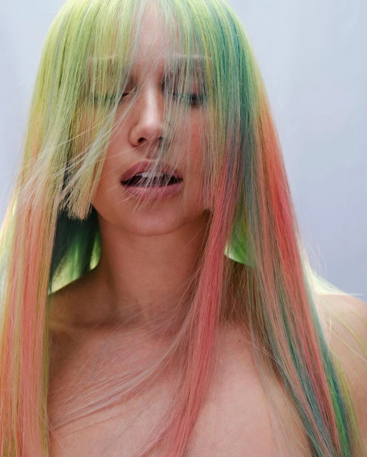 Close-up portrait of a woman with vibrant green, yellow, and pink dyed hair, eyes closed, in a bright studio setting.