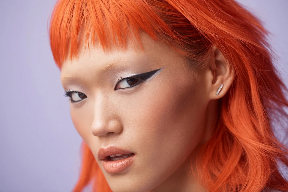 Close-up portrait of a young Asian adult female with orange hair, wearing heavy black eyeliner, against a light-purple background.