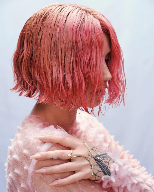 Profile view of a pink-haired person with fair skin, wearing a pink textured top, and adorned with gold bracelets against a light blue background.