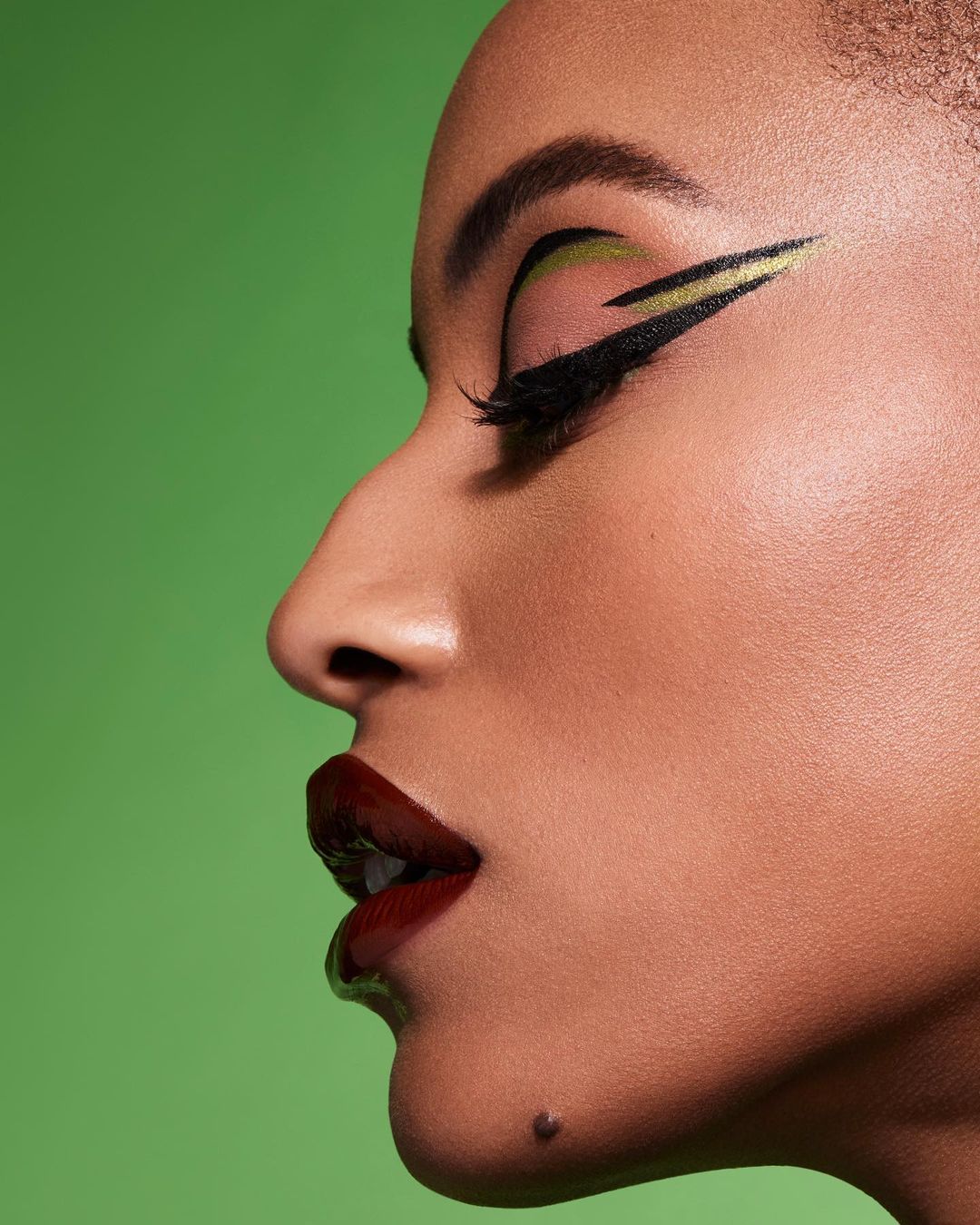 Close-up studio shot of a woman's face in profile with dramatic green and gold eye makeup, bold burgundy lipstick, and a green background.