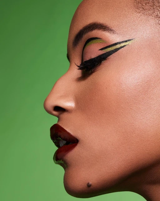 Close-up studio shot of a woman's face in profile with dramatic green and gold eye makeup, bold burgundy lipstick, and a green background.