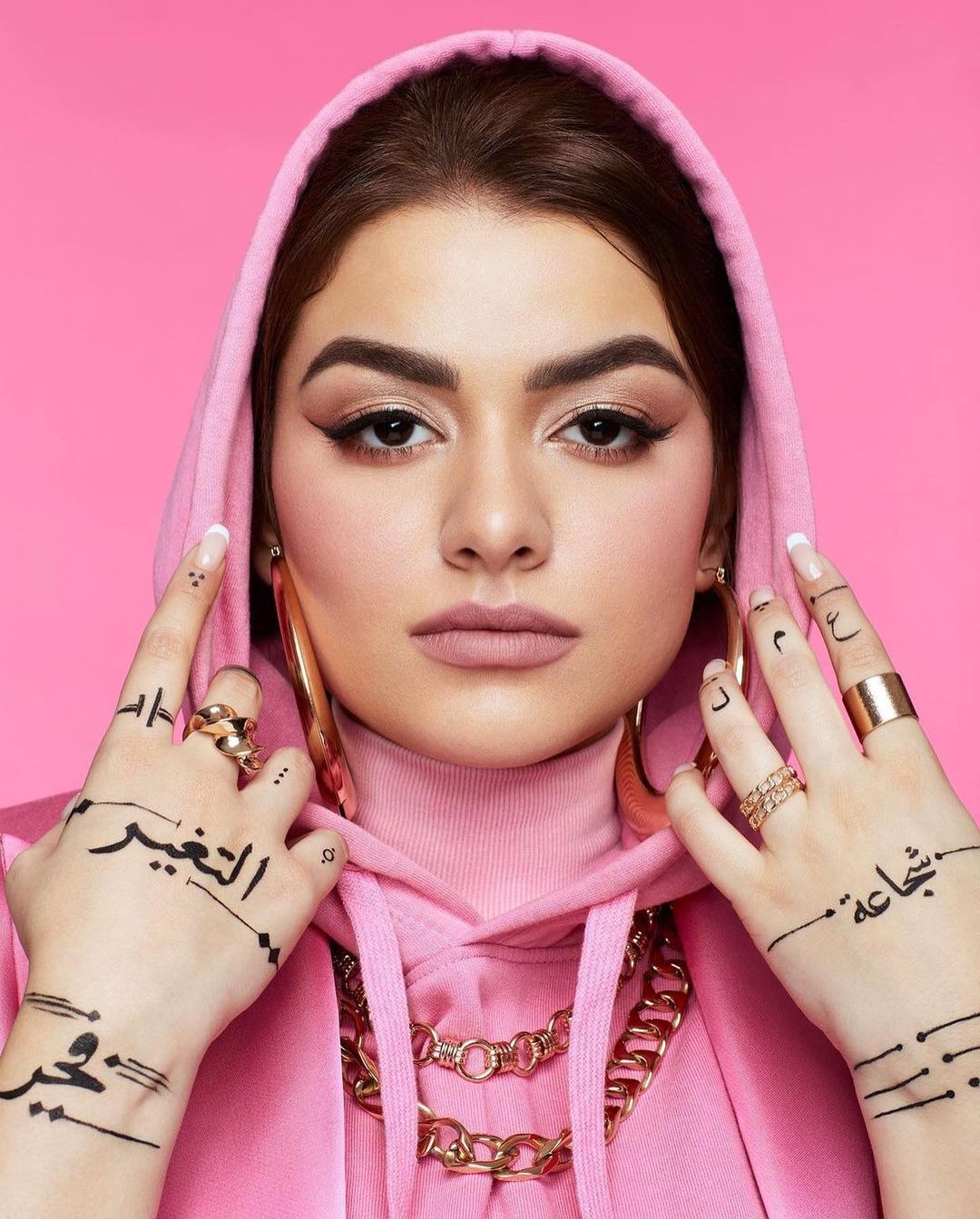 Portrait of woman with dark hair and pink hoodie, gold jewelry, face makeup, Arabic tattoos on her hands against pink backdrop.