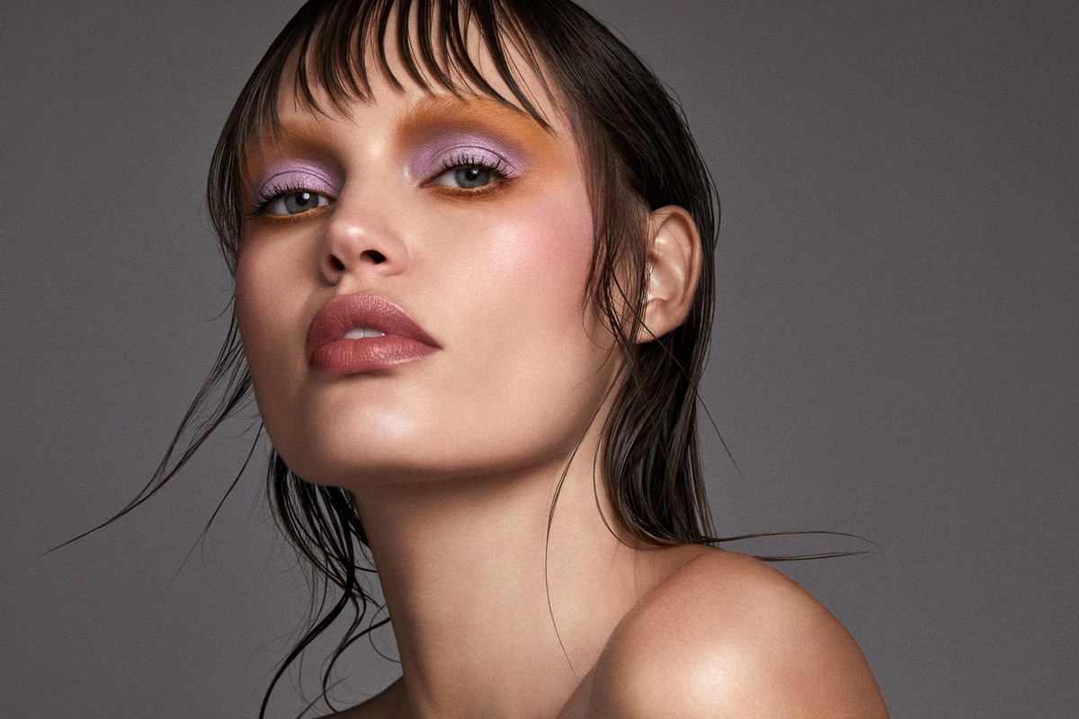 Close-up portrait of a model with colorful makeup, including lavender eyeshadow and glossy lips, set against a neutral gray backdrop.