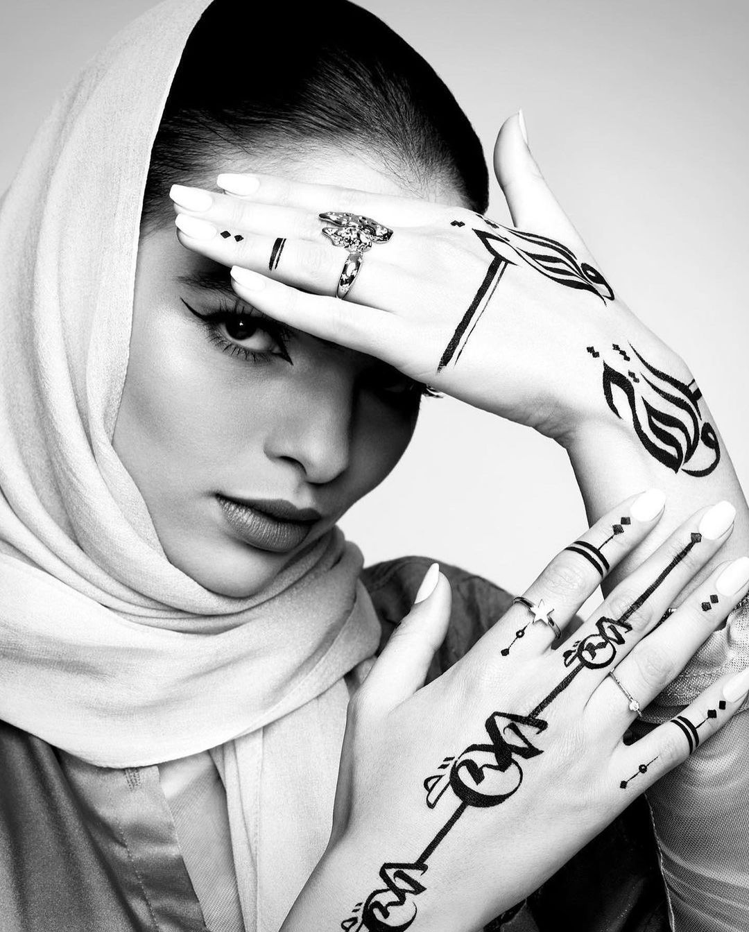 Black and white portrait of a woman with a hijab, looking at the camera, with Arabic script temporary tattoos on her hands.