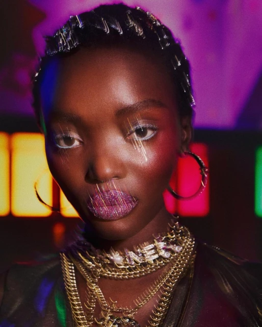Close-up portrait of an African American woman with glittery lips, white eyeliner, and layers of gold necklaces against a vibrant neon background.