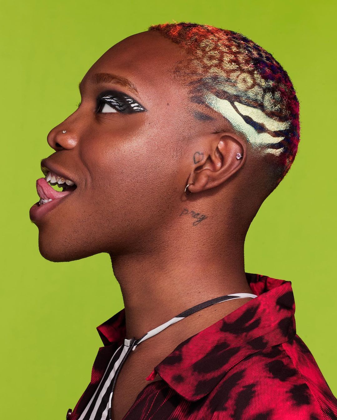 Close-up of person with colorful, patterned short hair, striking eye makeup, and tongue sticking out, posing against a lime green backdrop.