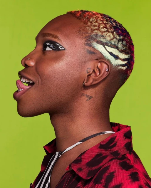 Close-up of person with colorful, patterned short hair, striking eye makeup, and tongue sticking out, posing against a lime green backdrop.