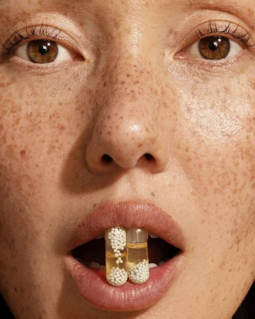 Close-up of a woman with freckles holding two clear capsules filled with liquid and small white balls between her teeth.