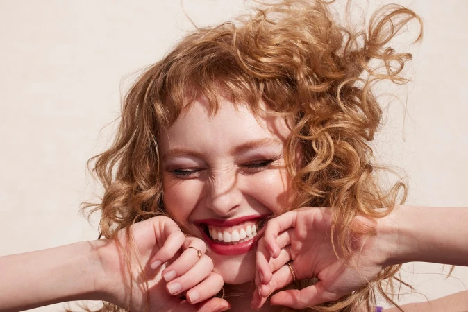 Close-up of a fair-skinned young woman with curly strawberry blonde hair, smiling broadly with hands framing her face outdoors.