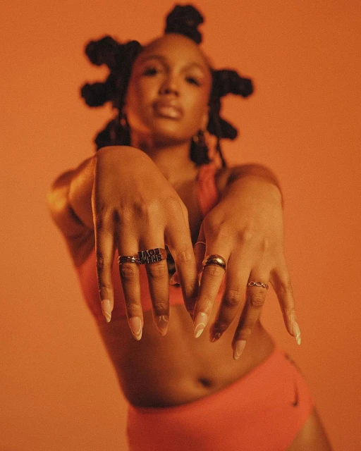 A woman poses in a coral sports bra and shorts against a matching background, showcasing rings on her fingers in a studio shot.