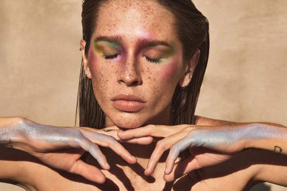 Close-up of a woman with colorful eye makeup and iridescent glitter on her hands, posing against a neutral-toned background in soft light.