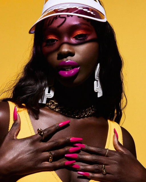 Portrait of a Black woman with vibrant makeup, hot pink nails, visor, and gold jewelry against a yellow background in a studio setting.