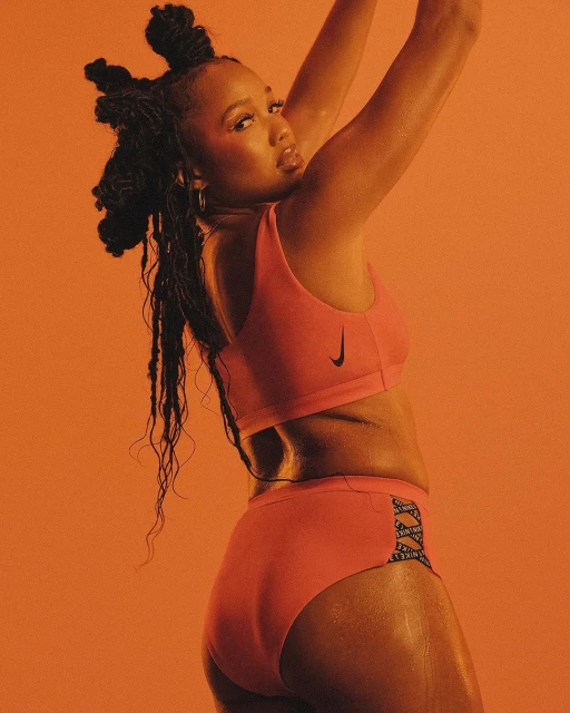 Athletic woman poses in pink Nike sports bra and high-waisted briefs against an orange backdrop, showcasing fitness and activewear.