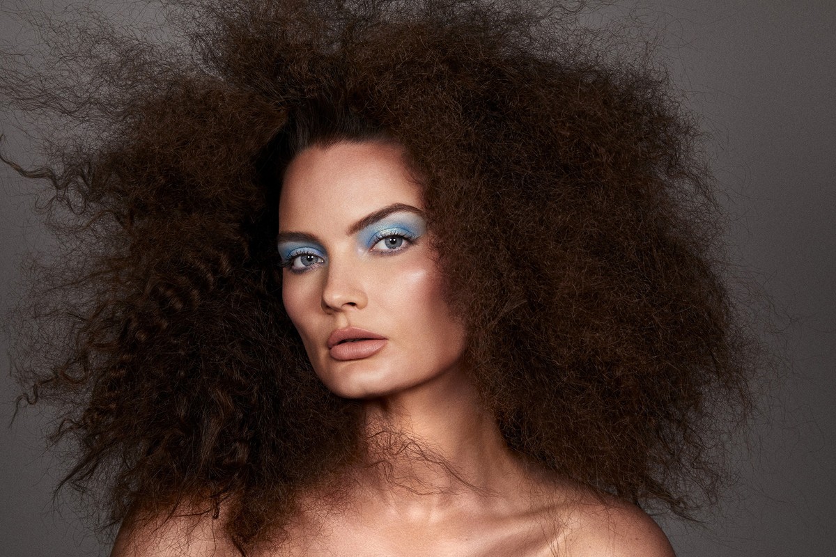 Portrait of a woman with large voluminous dark hair and bright blue eyeshadow photographed by Brooke Nipar against a grey background.