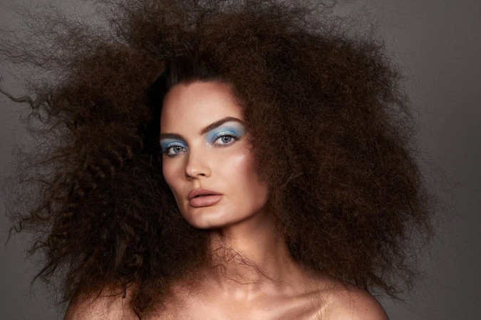 Portrait of a woman with large voluminous dark hair and bright blue eyeshadow photographed by Brooke Nipar against a grey background.