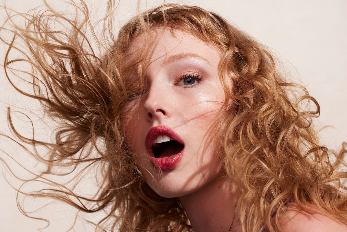 Close-up portrait of a beautiful young woman with curly strawberry blonde hair and red lipstick, photographed indoors against a beige background.