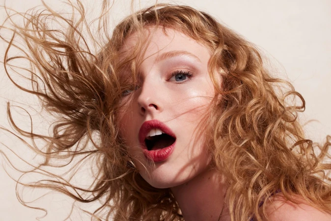 Close-up portrait of a beautiful young woman with curly strawberry blonde hair and red lipstick, photographed indoors against a beige background.