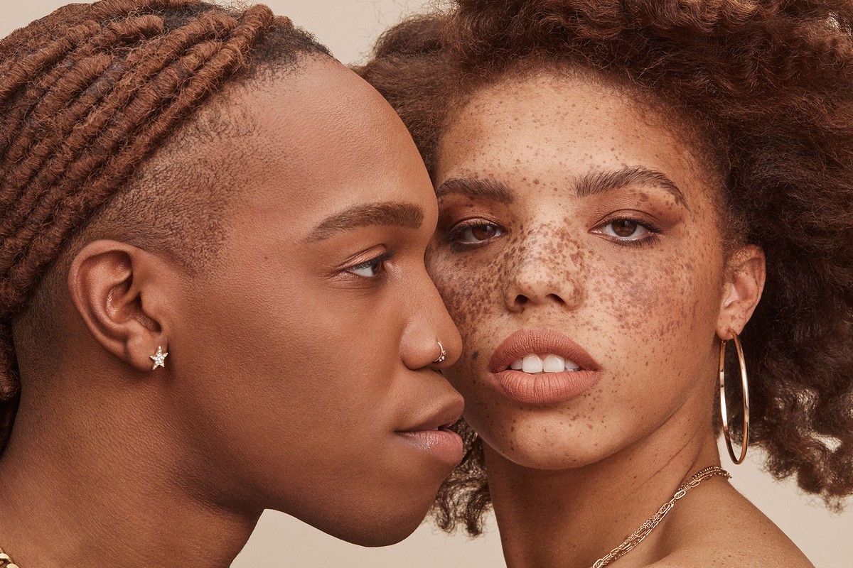 Close-up portrait of two models with warm skin tones, freckles, and natural hairstyles against a neutral background.