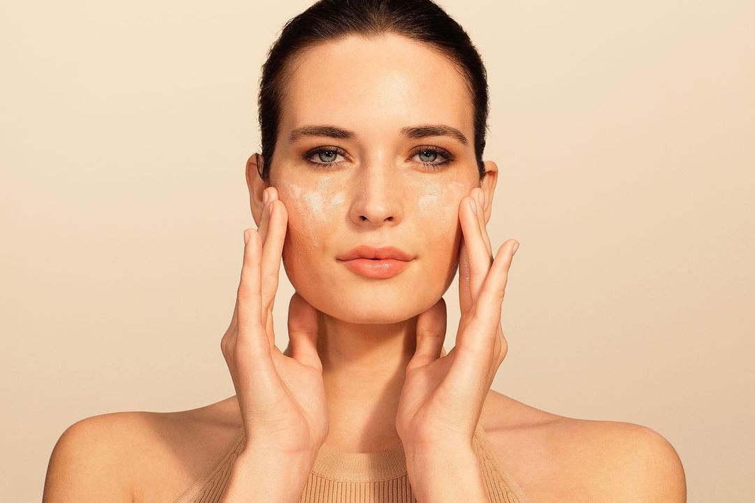 Close-up portrait of a woman applying skincare cream to her face against a warm neutral background.