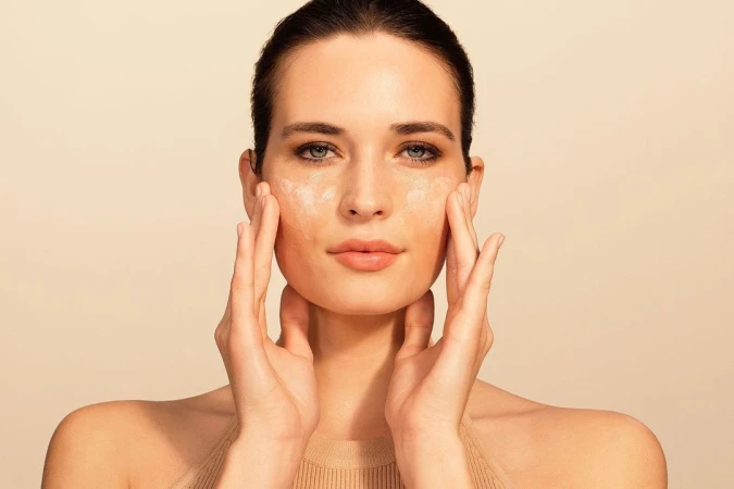 Close-up portrait of a woman applying skincare cream to her face against a warm neutral background.