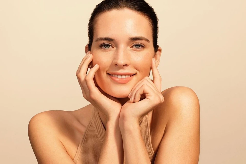 Smiling woman with her hands gently touching her face against a neutral background in a beauty headshot, creating a clean beauty aesthetic.