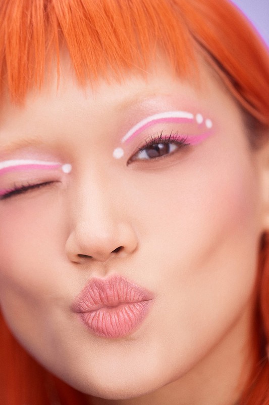 Close-up portrait of a young adult woman with orange hair winking, wearing pink and white makeup in an indoor environment.
