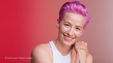 Smiling adult woman with short pink hair poses indoors against a gradient red and pink backdrop in a medium studio shot.