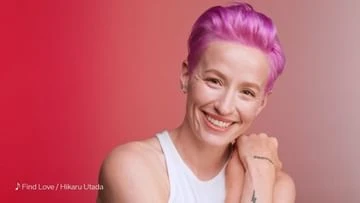 Smiling adult woman with short pink hair poses indoors against a gradient red and pink backdrop in a medium studio shot.
