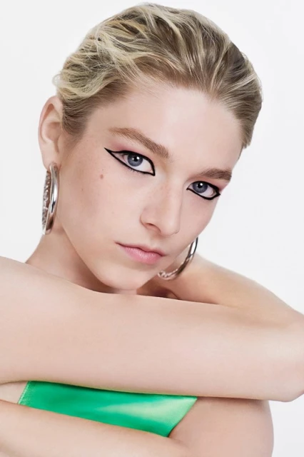Close-up portrait of a woman with graphic black eyeliner, silver hoop earrings, and a green satin top against a white background.