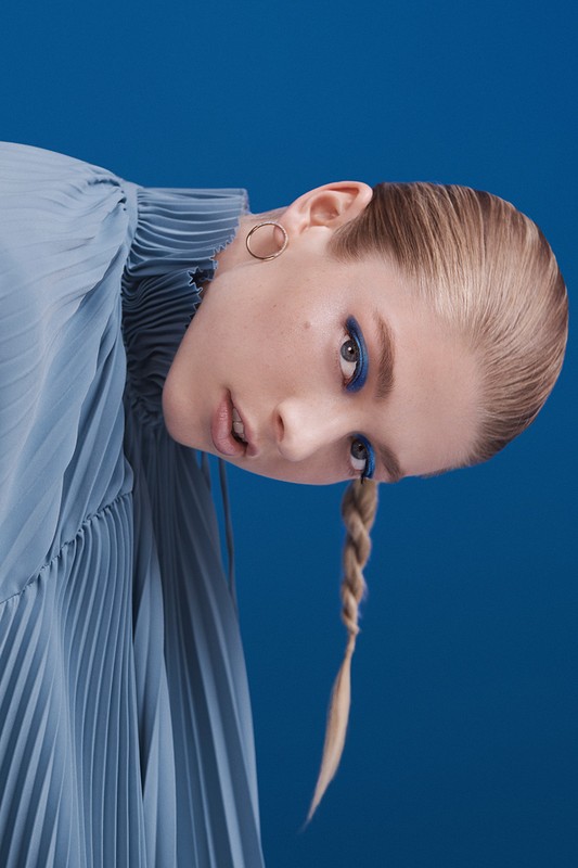 Portrait of a woman with blue eyeshadow and a braided ponytail wearing a light blue pleated top, set against a matching blue background.