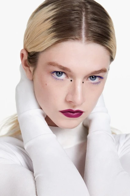 Striking beauty portrait of a blonde woman with purple makeup and white gloves, against a white backdrop in a clean studio shot.
