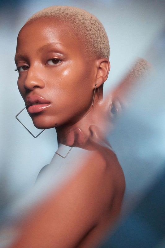 Close-up portrait of a woman with short blonde hair and square earrings against a soft blue gradient background.