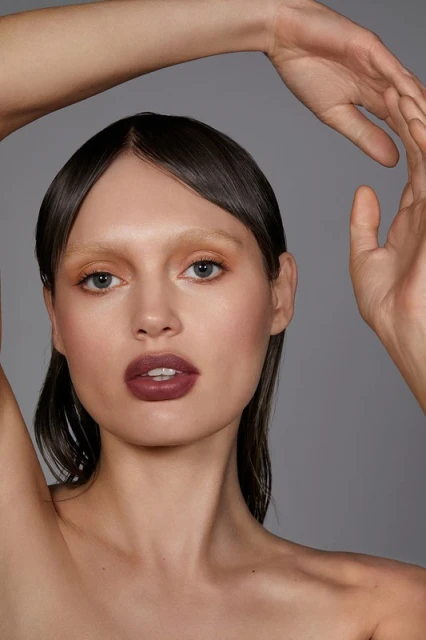 Close-up studio shot of a model with makeup, raising her hands in a beauty pose against a grey background, showcasing a modern aesthetic.