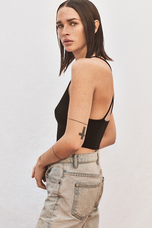 Portrait of a young adult female with shoulder-length brunette hair, wearing a black top and light-wash denim jeans against a white backdrop.