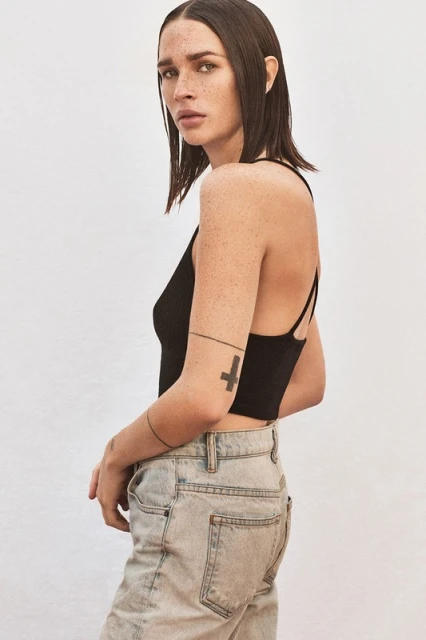 Portrait of a young adult female with shoulder-length brunette hair, wearing a black top and light-wash denim jeans against a white backdrop.