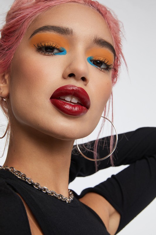 Close-up of a woman with pink hair and bold makeup featuring orange eyeshadow, blue underliner, and dark red lipstick, wearing a black top and jewelry.