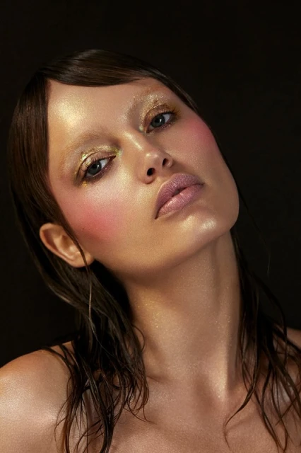 Close-up portrait of a woman with gold eyeshadow, blush, and glossy lips against a dark background in a beauty and fashion shot.