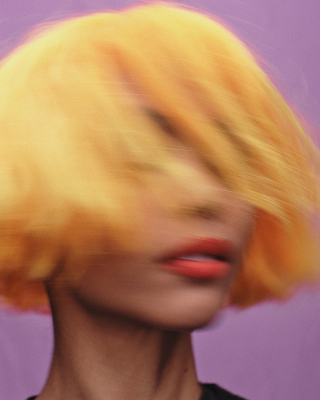 Close-up portrait of an adult female with orange short hair and coral lipstick, set against a solid purple backdrop, motion blurred.