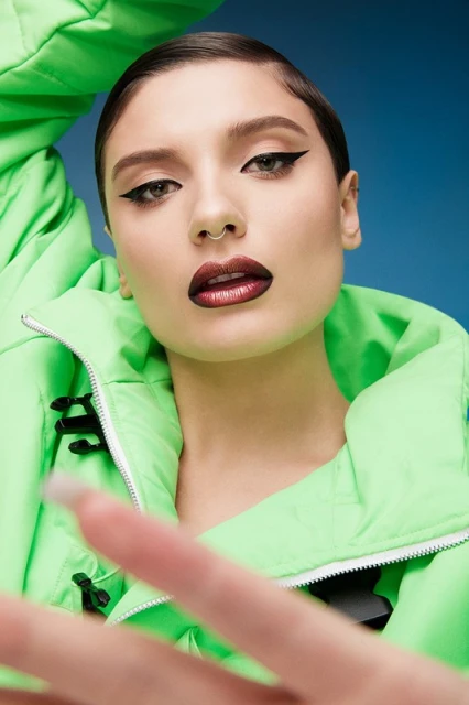 Close-up portrait of a young adult woman with dark lipstick and winged eyeliner, wearing a bright green puffy coat against a blue backdrop.