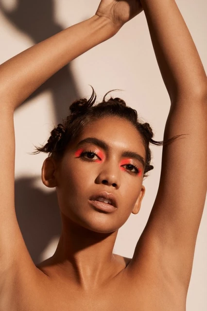 Close-up studio shot of a young adult woman with arms raised, wearing neon pink eye makeup, neutral lipstick and a playful hairstyle.