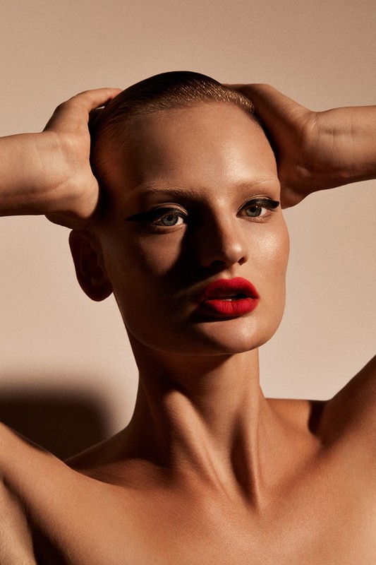 Close-up portrait of a woman with slicked-back hair, winged eyeliner, and vibrant red lipstick holding her hands behind her head.