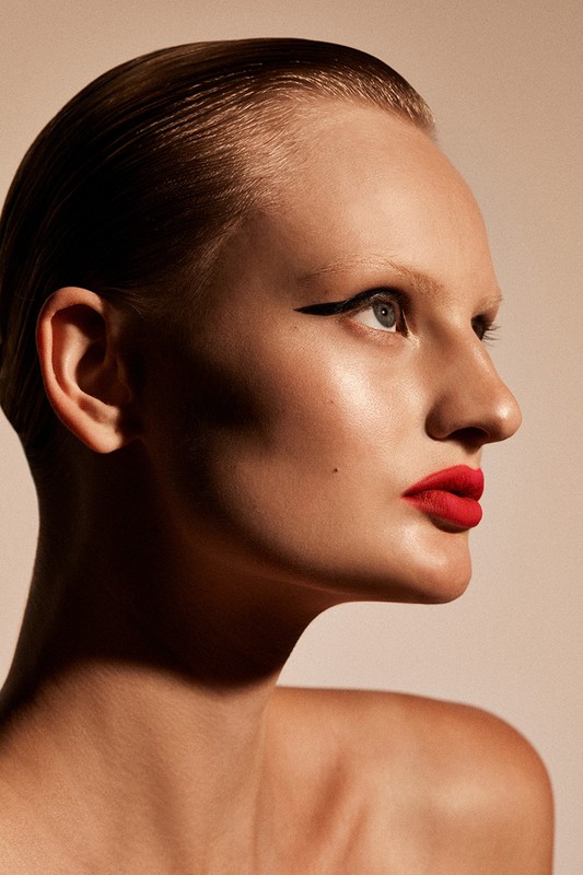 Close-up studio headshot of a young adult woman with red lipstick and sleek hair, gazing upwards against a neutral background.