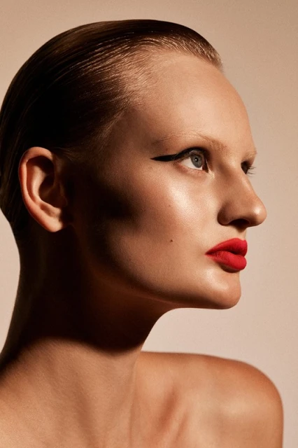Close-up studio headshot of a young adult woman with red lipstick and sleek hair, gazing upwards against a neutral background.