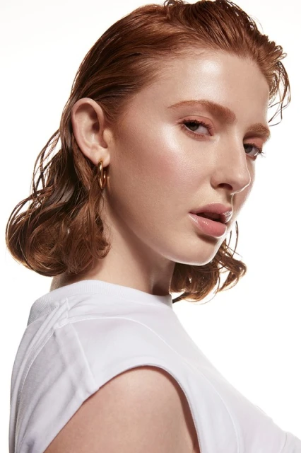 Close-up portrait of a fair-skinned, adult female with wavy red hair and gold hoop earrings, wearing a white t-shirt against a white backdrop.